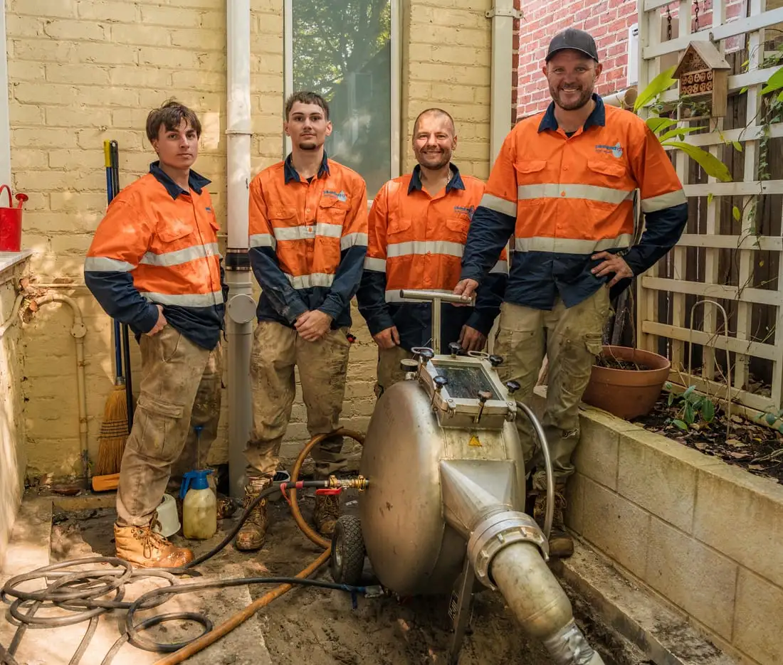 Four of the relining crew standing with the inversion drum in the side yard of a cream brick character home, the excavated access hole and the cut sewer pipe open in the sand in front of them.