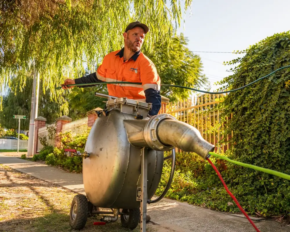A relining technician standing behind a stainless steel inversion drum on the verge of a Perth home, feeding the green liner hose out towards the drain access point.