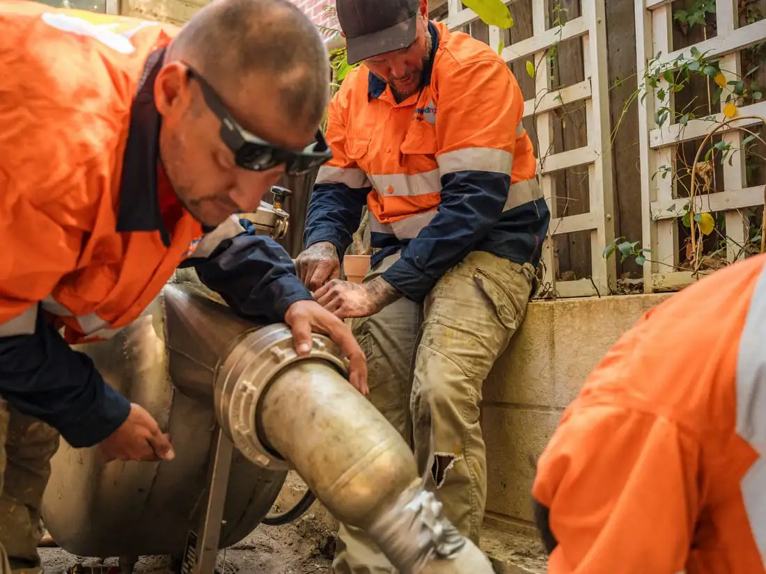 Three of the crew crouched in a narrow side passage of a character home, guiding the liner out of the inversion drum and into the open end of the sewer pipe at the bottom of a hand-dug access hole.