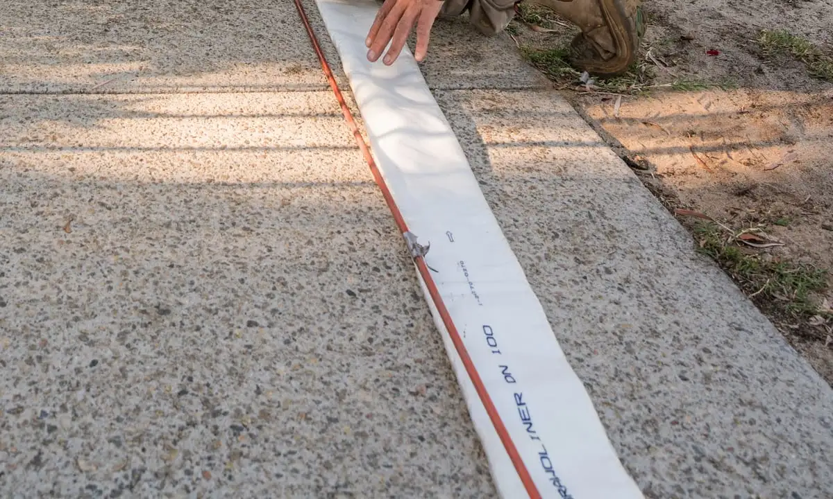 A flat, uninflated relining liner laid out along a concrete footpath before it goes in, printed with its diameter marking, with a technician's hand flattening it and the orange draw rope running the length of it.